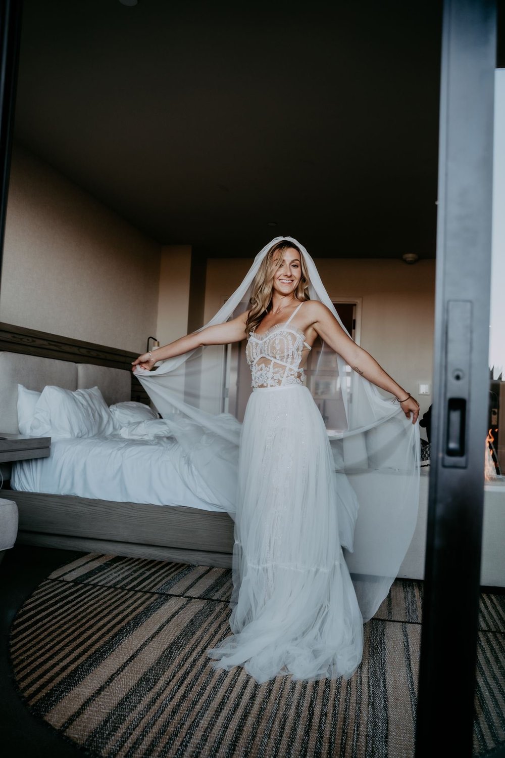 Bride in a lace sheath gown lifting a blusher veil away from her face in a softly lit room