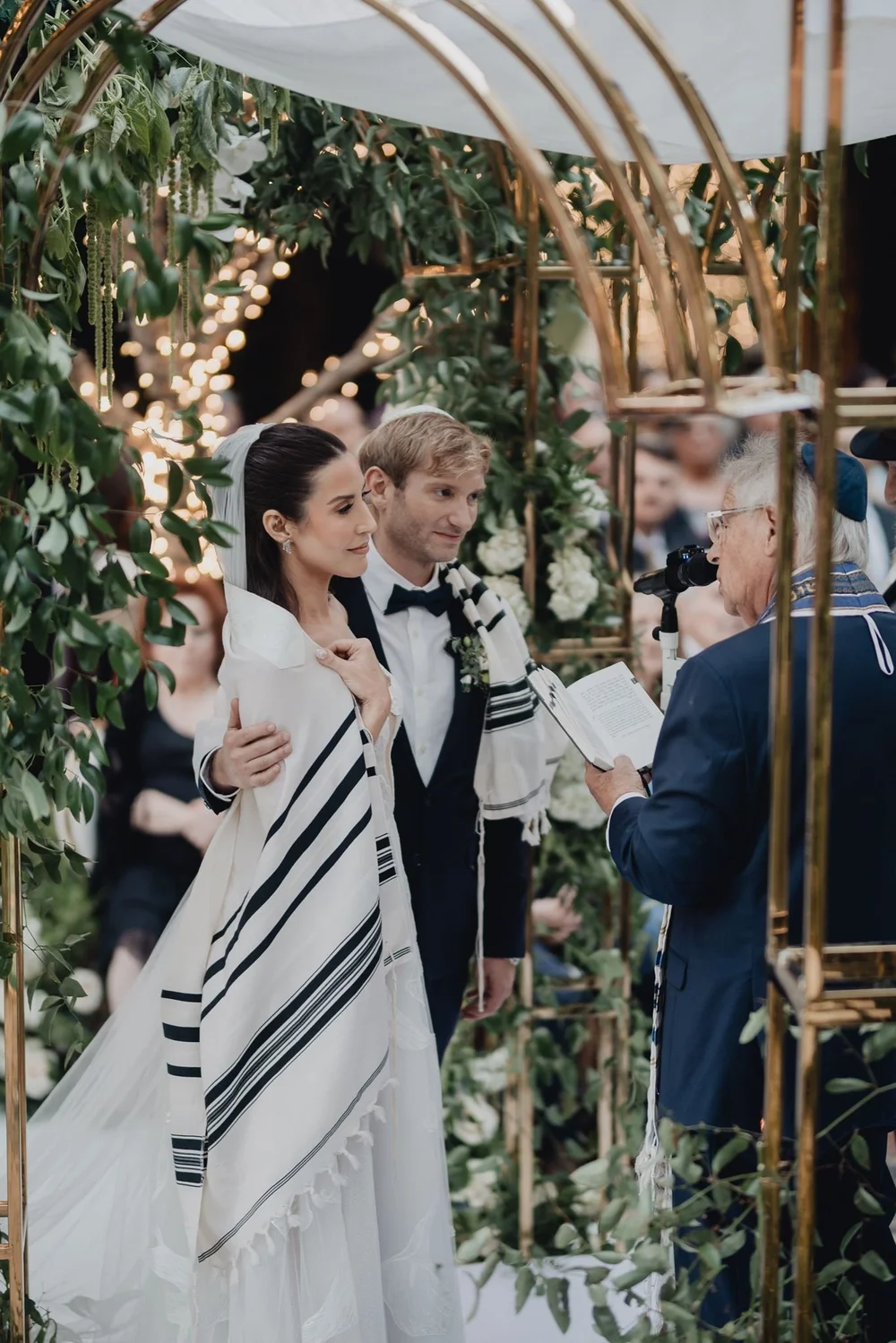 Bride in a striped tallit beside the groom under a gold and greenery chuppah