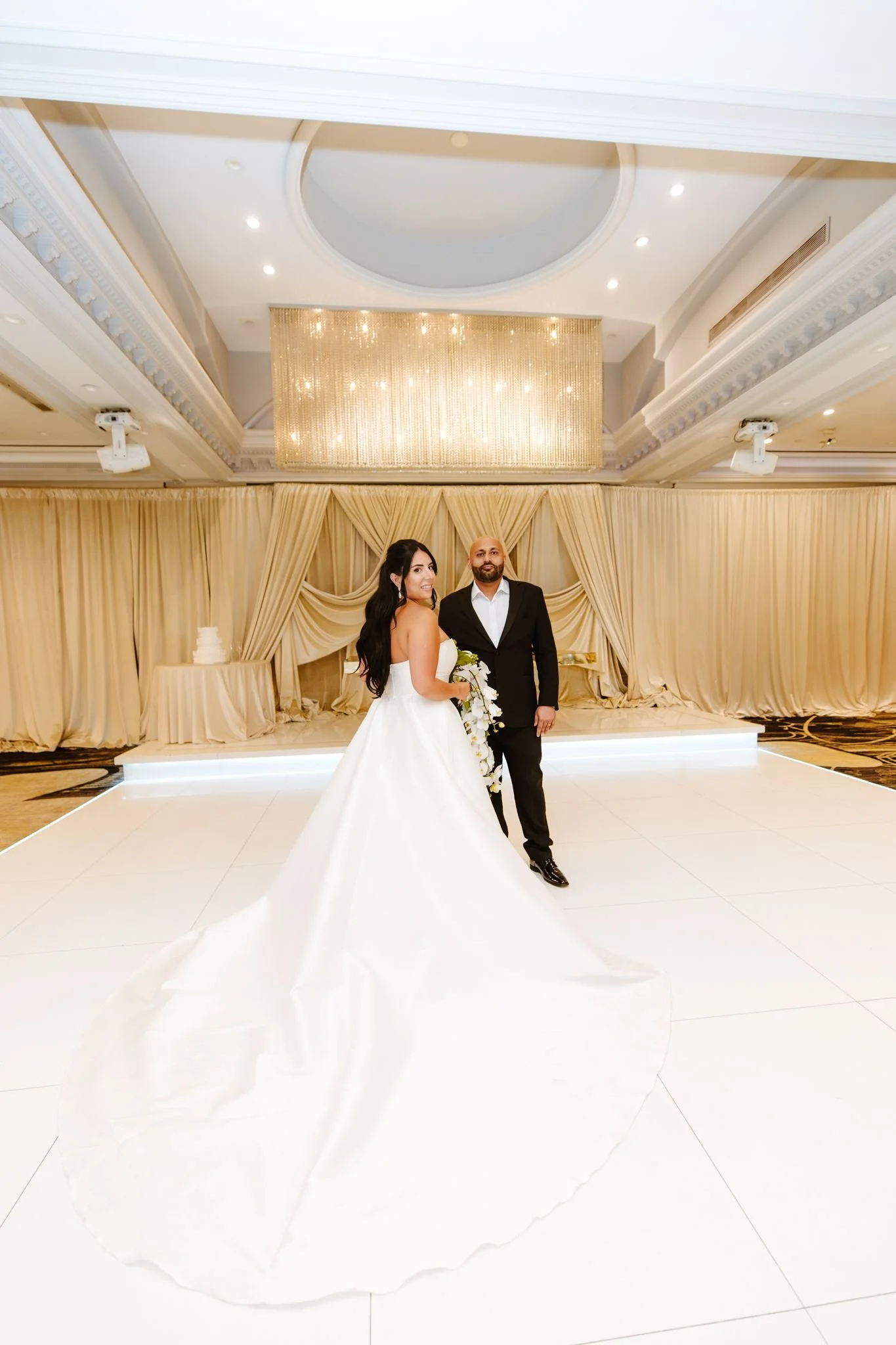 Bride and groom under the domed ceiling and crystal light panel at Glenoaks Ballroom, her train across the floor
