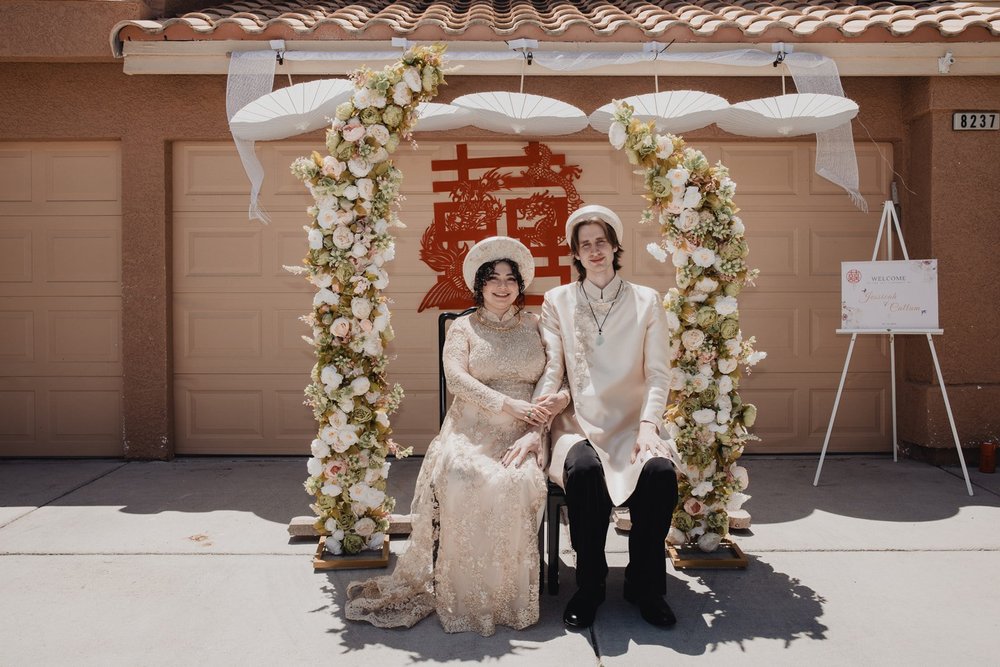 Couple in cream ao dai and khan dong seated before a floral arch and double-happiness sign at their Vietnamese wedding