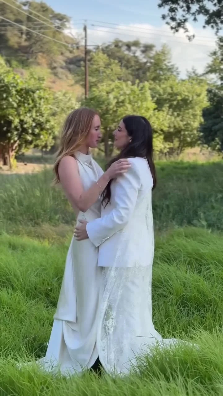 Two brides holding each other in tall grass under an open sky, filmed vertically on a phone
