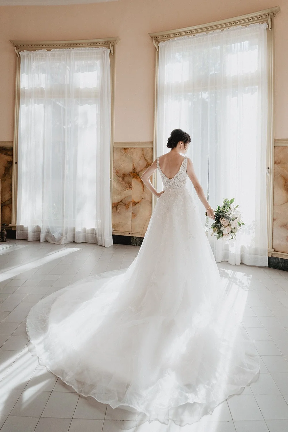 Bride in a tulle A-line gown with a wide train fanned out in a sunlit room with tall curtained windows