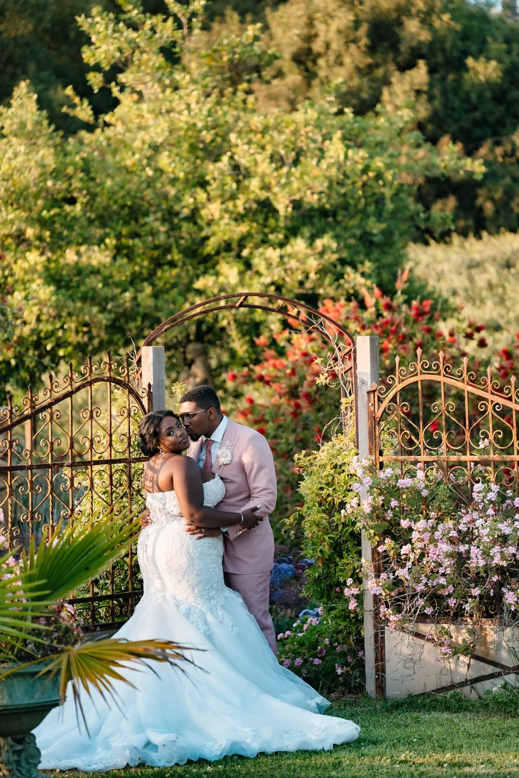 Bride and groom beside an ornate iron garden gate surrounded by flowering shrubs at Ethereal Gardens in Escondido