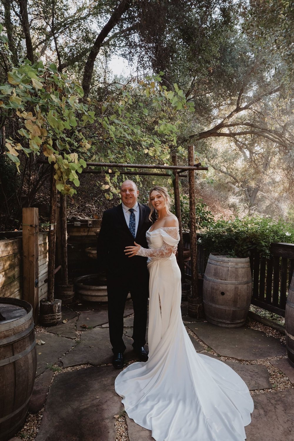 Bride in an off-the-shoulder silk sheath gown with a sweep train walking a stone courtyard with her father