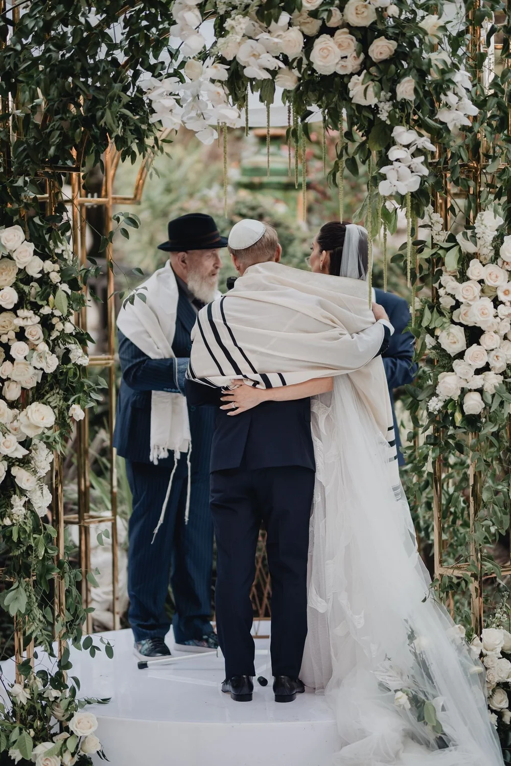 Couple wrapped together in a tallit beneath a white-rose chuppah