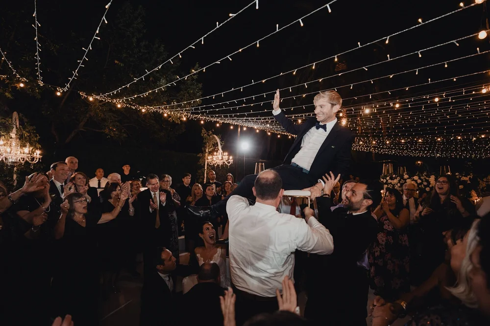 Groom lifted on a chair during the hora under strands of string lights