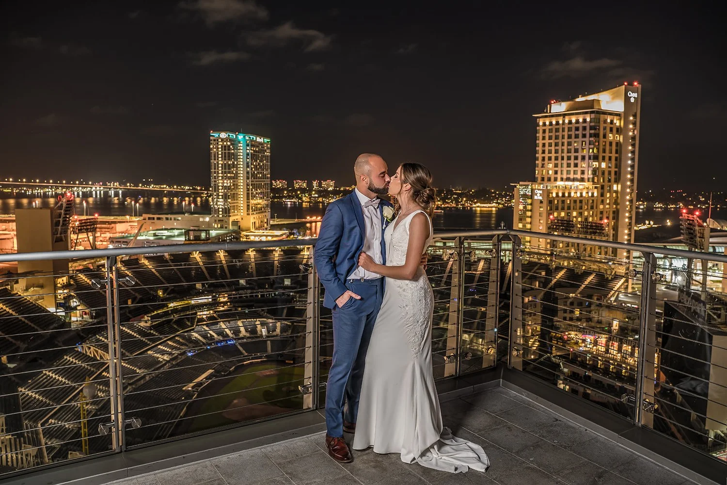 Couple on the balcony of The Ultimate Skybox at night with Petco Park below and the San Diego waterfront lit behind them