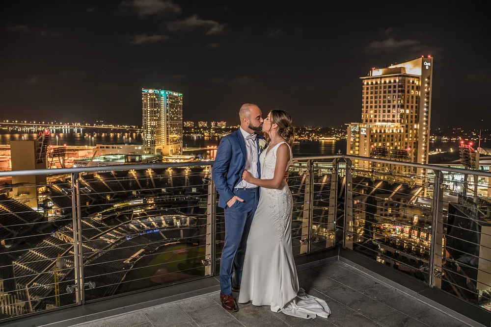 Couple on the balcony of The Ultimate Skybox at night with Petco Park below and the San Diego waterfront behind