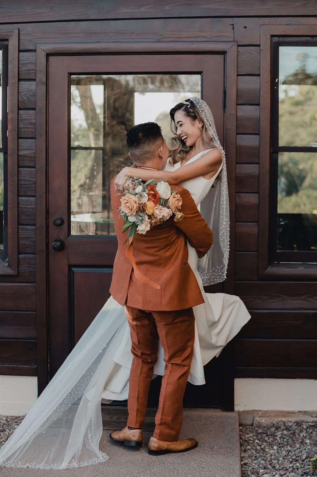 Bride and groom beside the heavy dark timber doorway at Mt. Woodson Castle, the groom in a rust-coloured suit