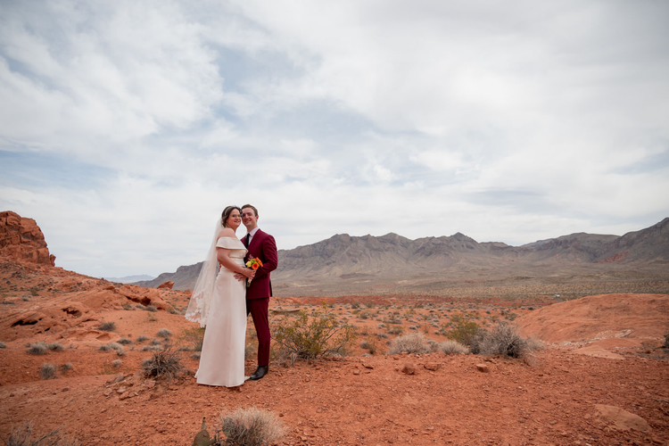 Bride in an off-the-shoulder gown and groom in a burgundy suit stand on red rock with the desert mountains stretching out behind them at Valley of Fire.