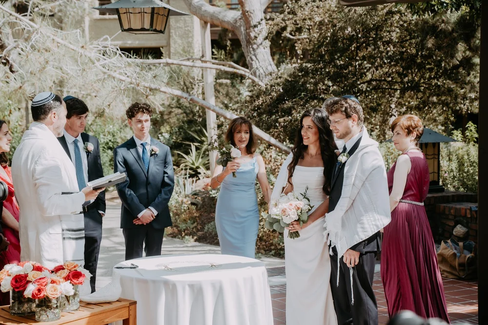 Wedding party gathered outdoors around the signing table while a guest in a tallit reads the text