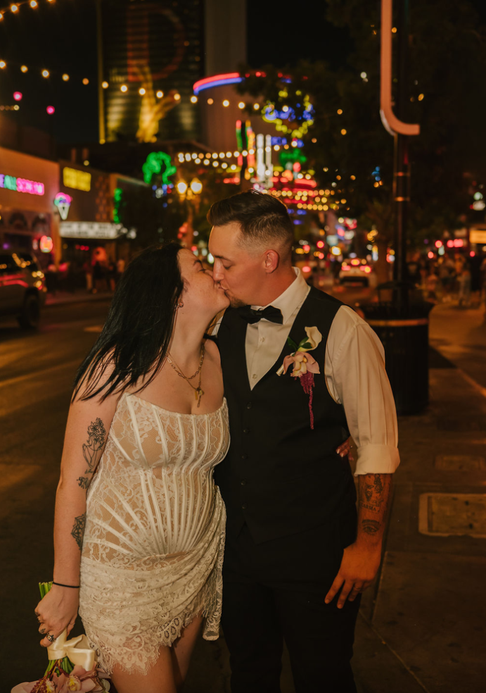 Couple kiss on Fremont Street at night under neon signs and string lights, the bride in a short lace corset dress holding her bouquet.