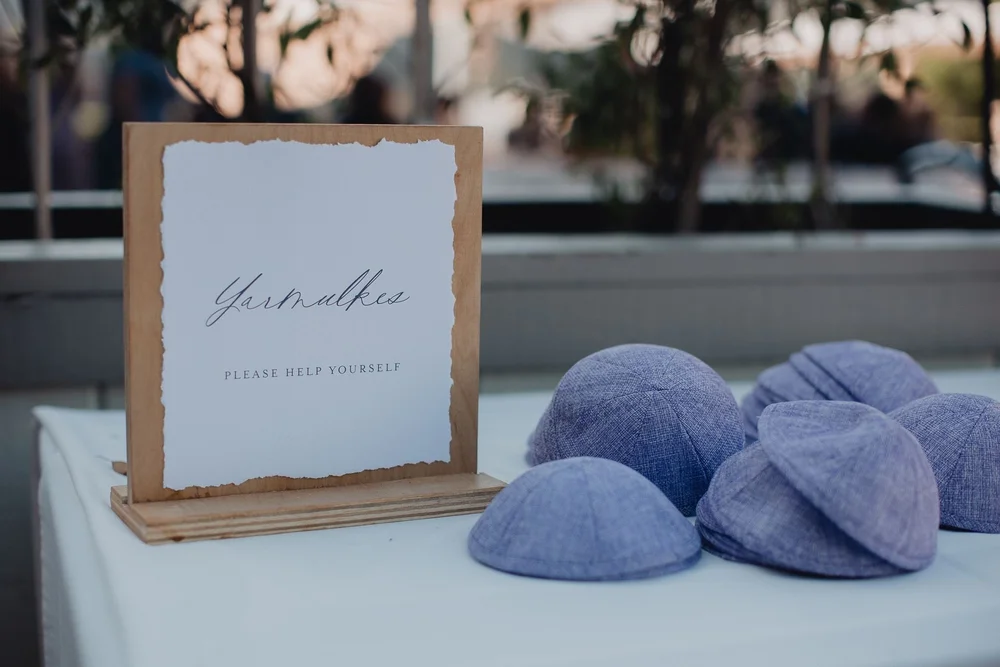 Tray of blue kippot beside a hand-lettered sign inviting guests to help themselves