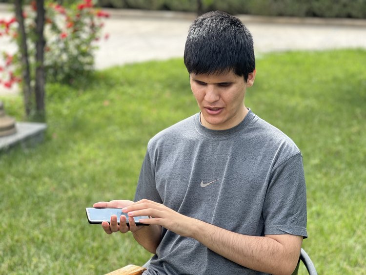 Photo of a young man sitting in a grassy area, his face facing ahead and away from the phone that he is holding in his right hand. With his left hand, he is touching the screen.