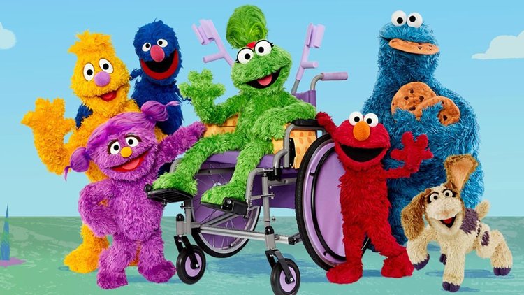 Photograph of seven of the muppets including the green muppet, Ameera in her ill fitting wheelchair