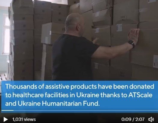 A screen shot of WHO Ukraine film. A man stands in front of boxes of Assistive Products. Caption reads, 'Thousands of assistive products have been donated to healthcare facilities in Ukraine thanks to ATscale and Ukraine Humanitarian Fund