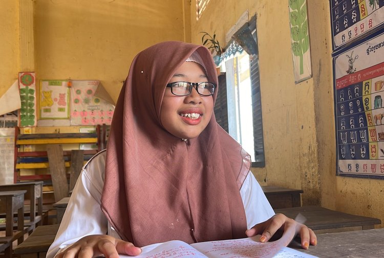A girl is sitting in a classroom in Cambodia. She is smiling and wearing glasses and has a book open in front of her