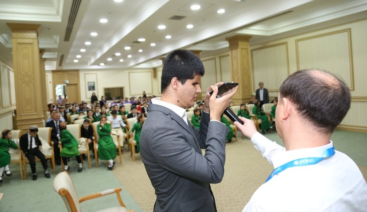 A young man, who is blind, is demonstrating how to use the new TTS technology at the event. He is holding a phone in front of the audience. There is another man next to him who is holding a microphone.
