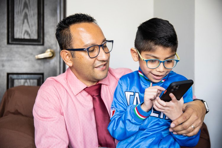 Father wearing glasses, a pink shirt and maroon tie, and young son wearing glasses, a blue jersey, sitting on his father’s lap using a mobile phone, both the father and son are engaged in what the son is doing. Looks like he is pointing to something