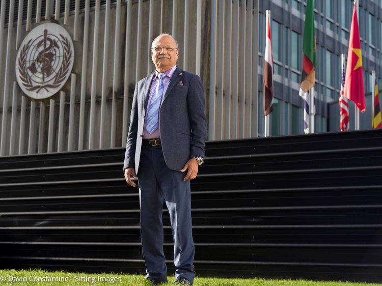 Former Head of AT at WHO, Chapal Khasnabis wears a suit and is standing in front of some steps outside and in front of the WHO building in Geneva