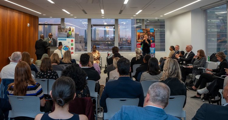 A room is full of people who are all sitting down, with their backs to the camera, looking at the speakers. In the background are two UNICEF posters, a podium and one of the speakers, standing in front of a window. Through the window, UNHQ is seen.
