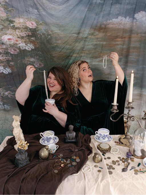 Louisa and Annie sit in velvet gowns, looking at the camera, as they eat jewelry off of fine silver