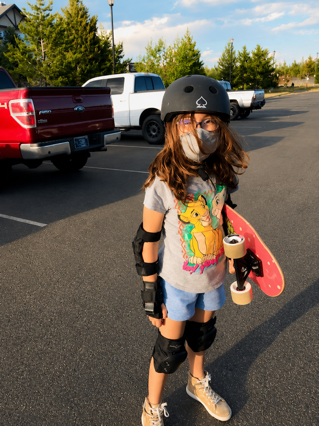 A child wearing a helmet, face covering, knee pads, and elbow pads holds a red skateboard in a parking lot.