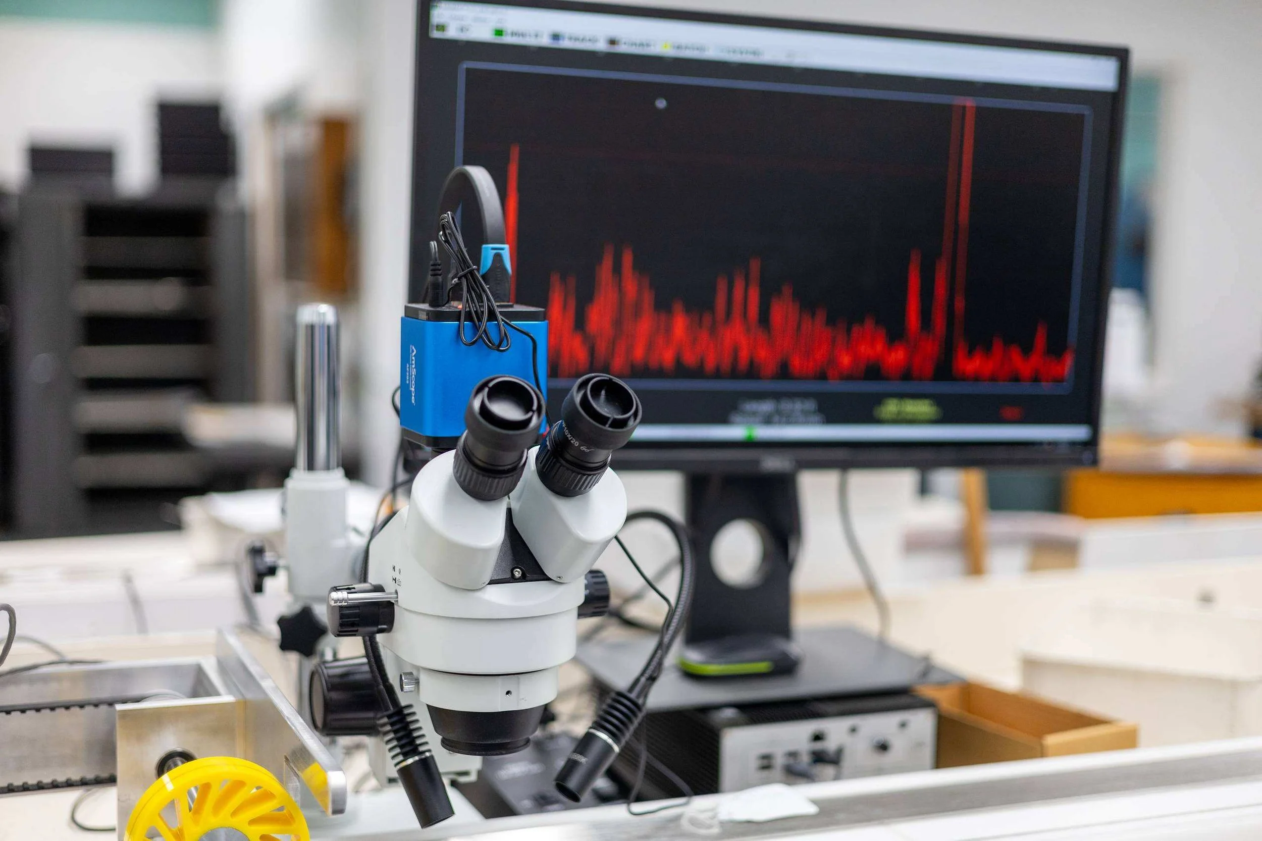 Stereo microscope on an inspection bench with elemental analysis output displayed on the monitor behind