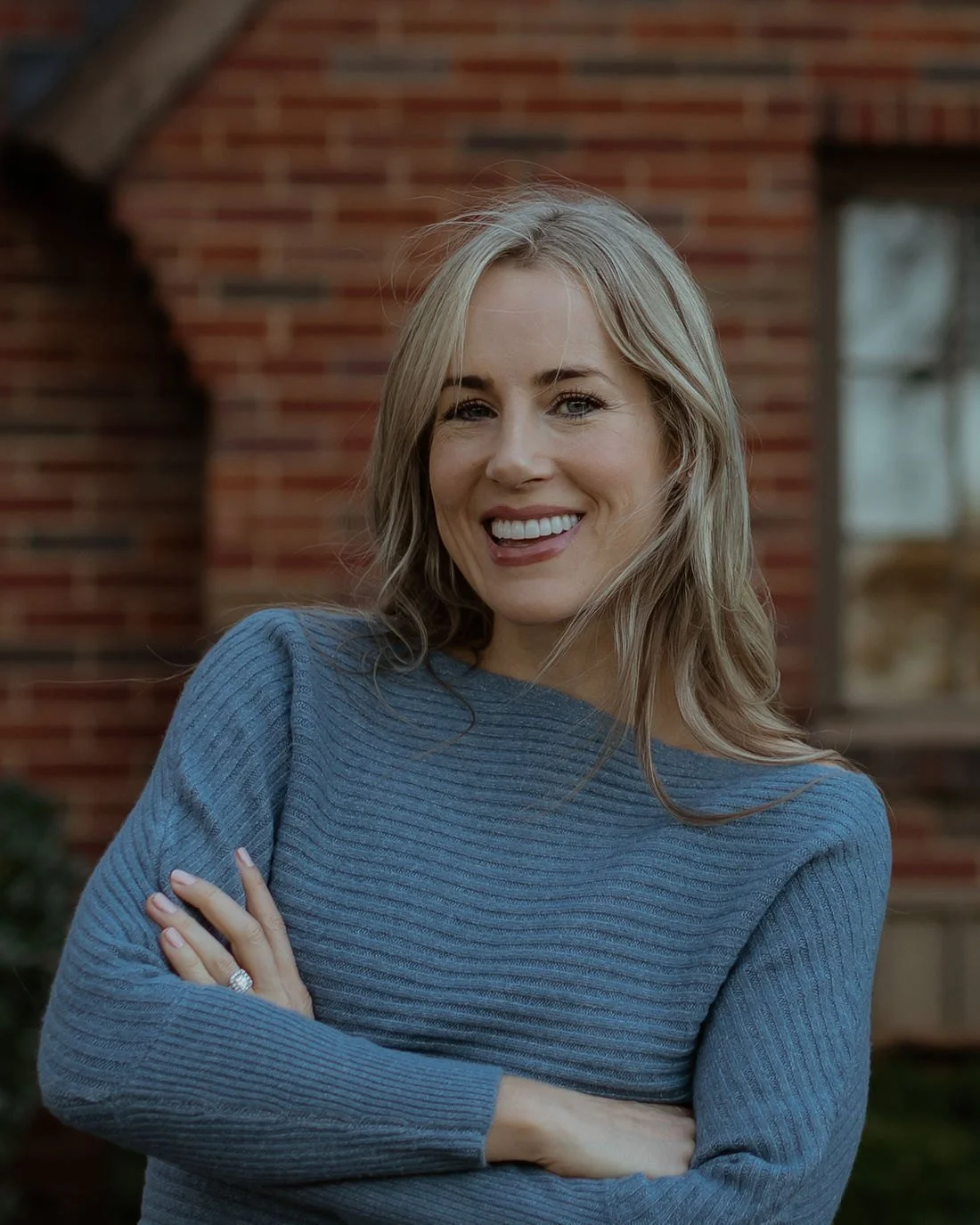 Megan Bruneau in a blue knit dress, arms crossed, smiling in front of a brick house