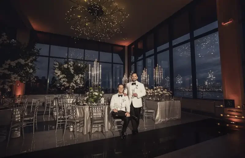 A couple in white dinner jackets in a candlelit reception room with tall floral centerpieces and a city skyline view