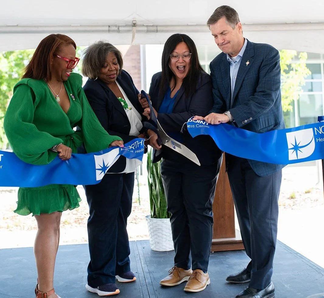 Four people cut a blue NorthPoint Health & Wellness Center ribbon with oversized scissors under a white tent. From left: NorthPoint Community Board Chair Ticia Selmon, NorthPoint CEO Stella Whitney-West, Hennepin County Commissioner and Board Chair Irene Fernando, and former Hennepin County Administrator David Hough.