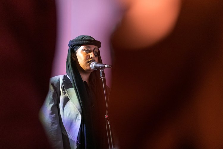artist asmaa jama wearing a black headscarf and jacket, speaking into a microphone, semi obscured by a red shadow