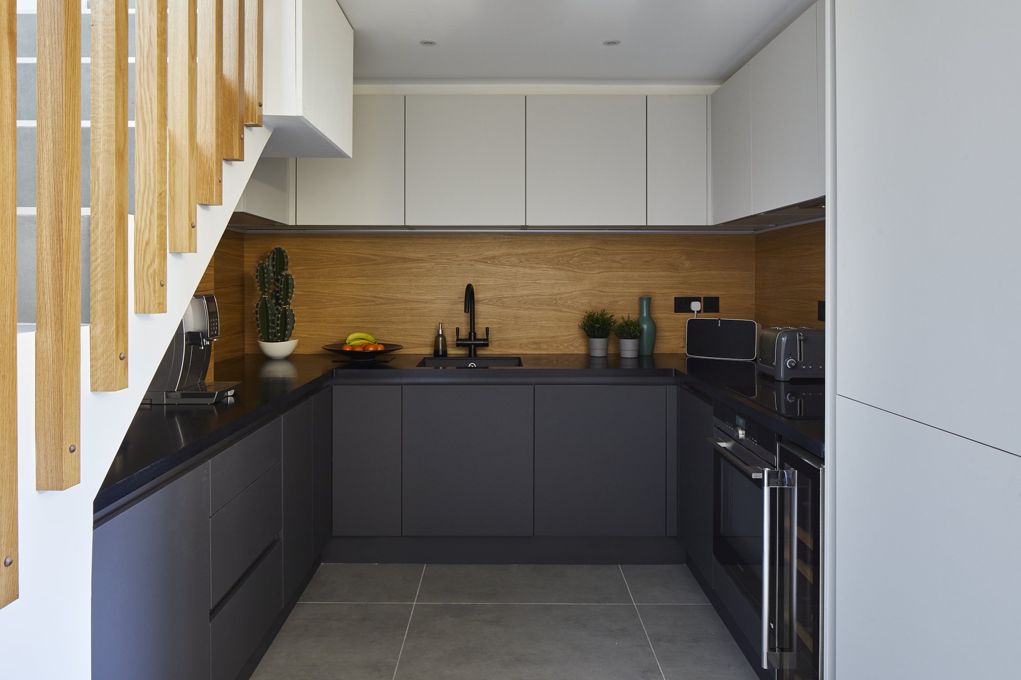 After photo — U-shaped industrial kitchen in a Shoreditch loft in daylight, showing matte black handleless lower cabinets, oak wood splashback, pale grey upper units and integrated appliances. Interior design by Studio Akiva
