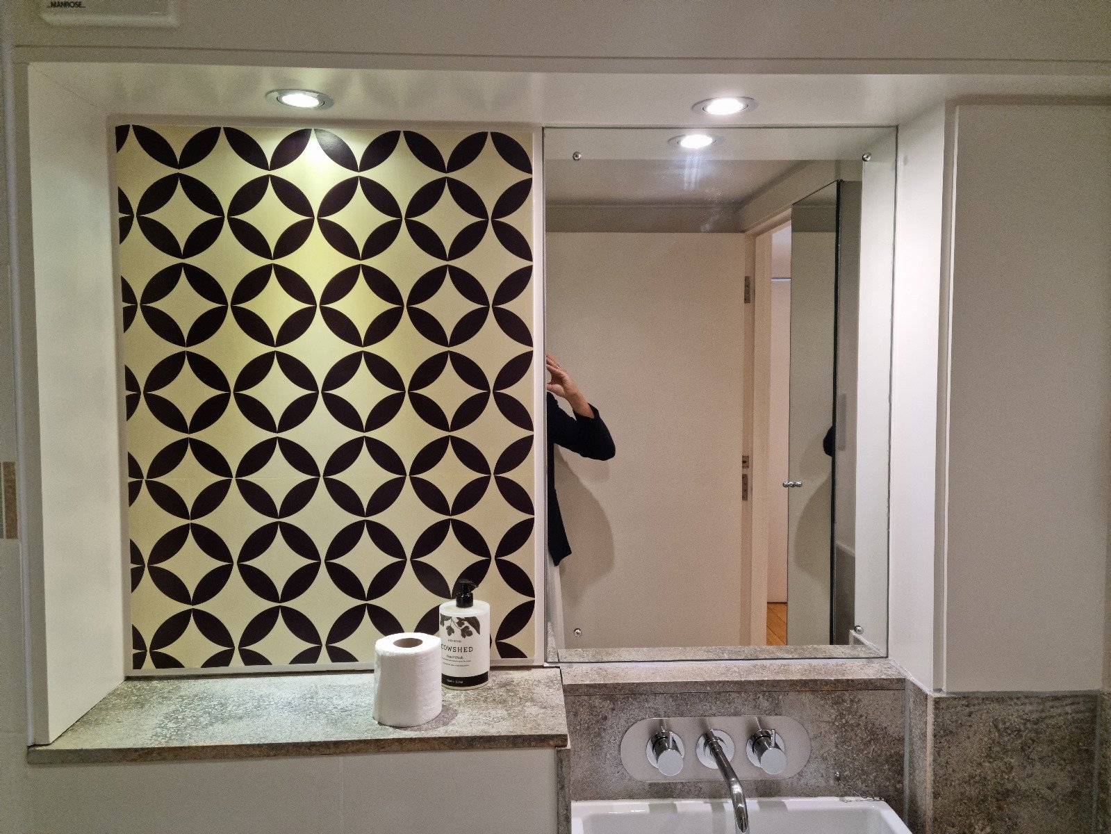 Before photo — bathroom in a Grade II listed school conversion duplex London, showing bold geometric patterned wallpaper panel, chrome taps, granite worktop and mismatched white tiles, prior to Studio Akiva redesign