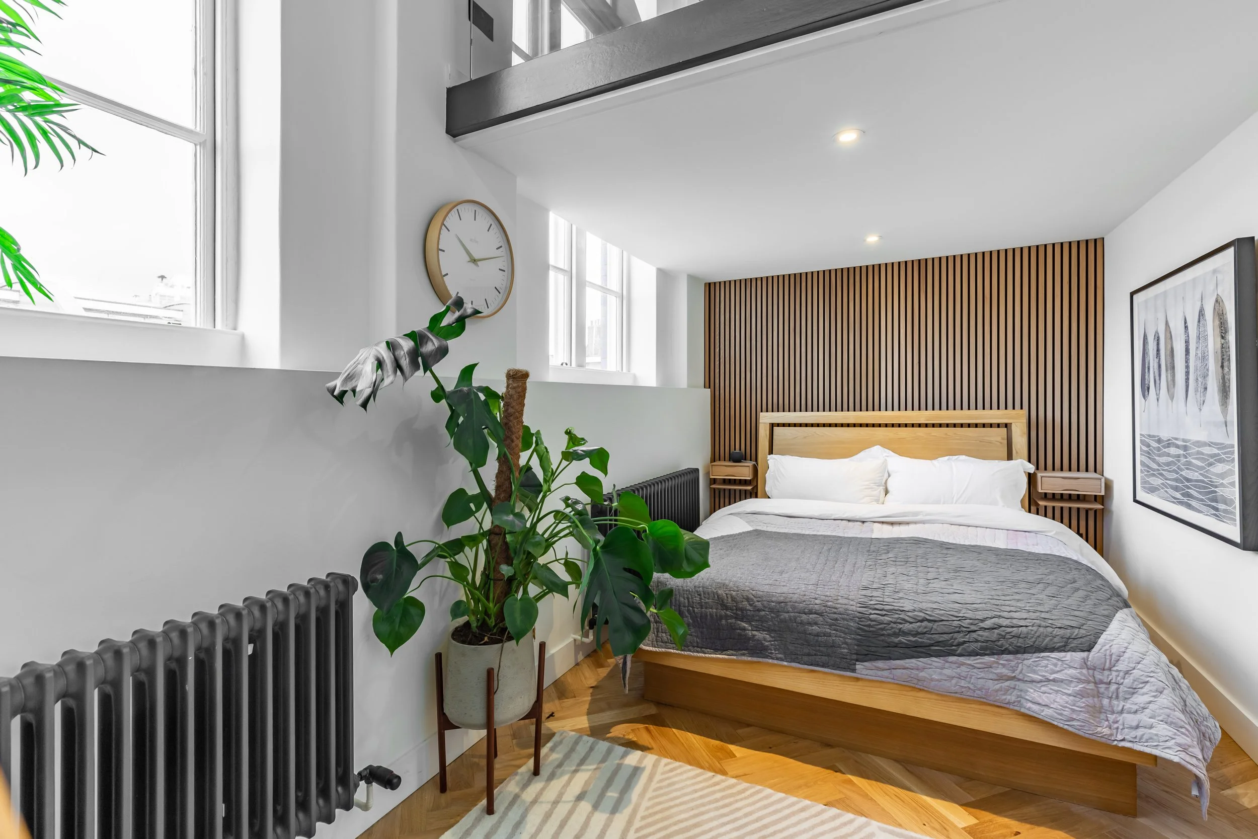 Master bedroom in a Grade II listed school conversion duplex London — full-width oak slat feature wall, oak bedframe with integrated side tables, herringbone parquet floor, matte black column radiators, large sash windows. Interior design by Studio Akiva