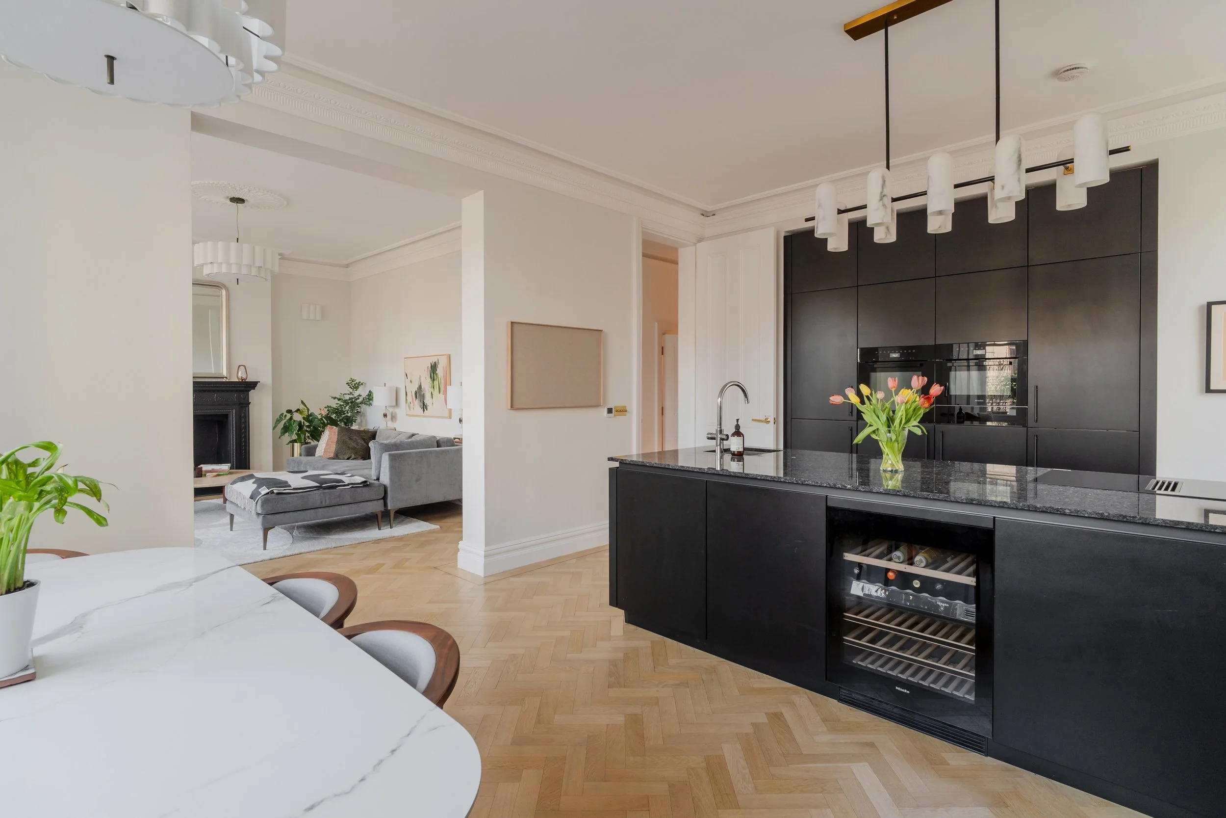 Contemporary black kitchen with marble island, herringbone flooring, and open-plan connection to elegant living space.