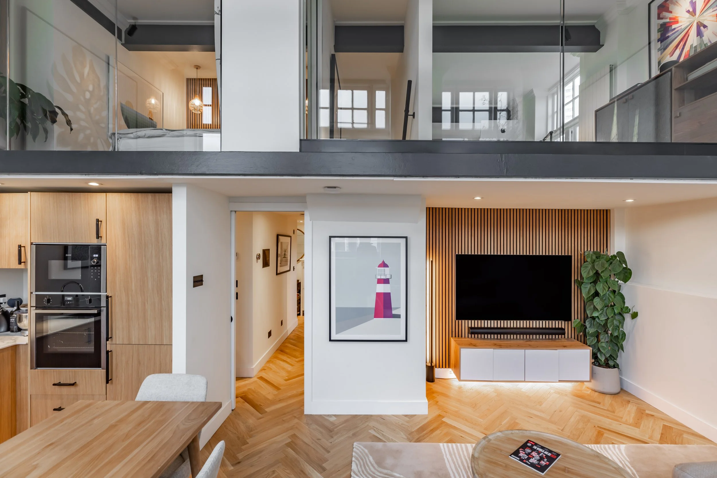 Aerial view of a Grade II listed school conversion duplex in London — oak kitchen cabinets below, mezzanine master bedroom visible through glass balustrade above, oak slat TV wall, herringbone oak floor, open-plan living and dining. Interior design by Studio Akiva