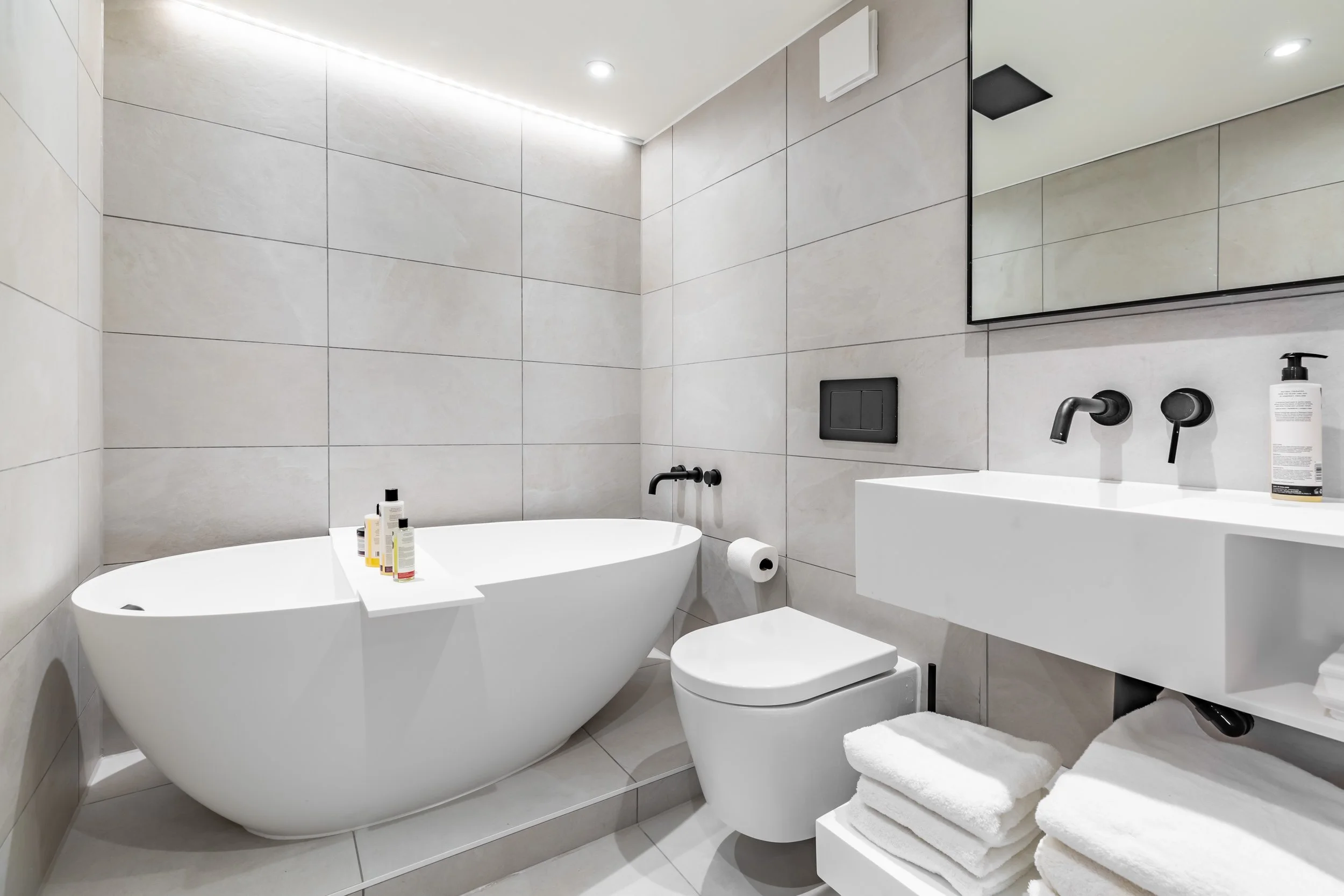 Main bathroom in a Grade II listed London duplex — freestanding white oval bath on stepped plinth, large-format pale grey tiles, matte black wall-mounted taps, black-framed mirror cabinet, LED ceiling strip. Interior design by Studio Akiva