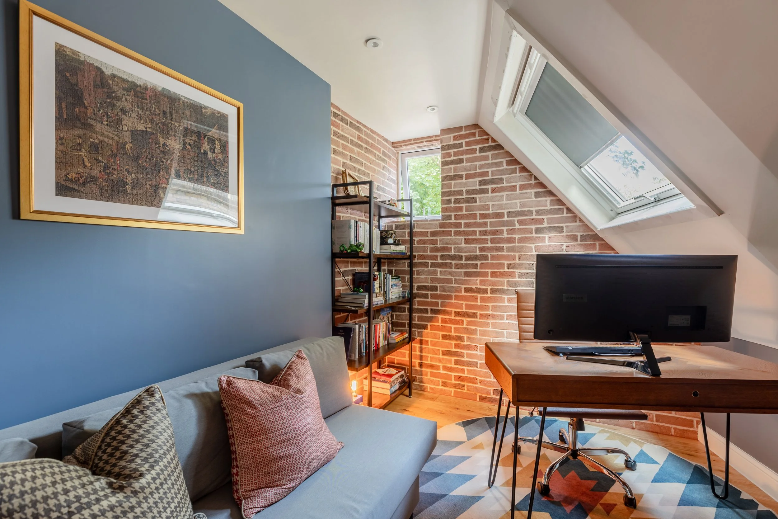 Modern loft home office with exposed brick walls, skylight window, blue feature wall, and mid-century style desk.