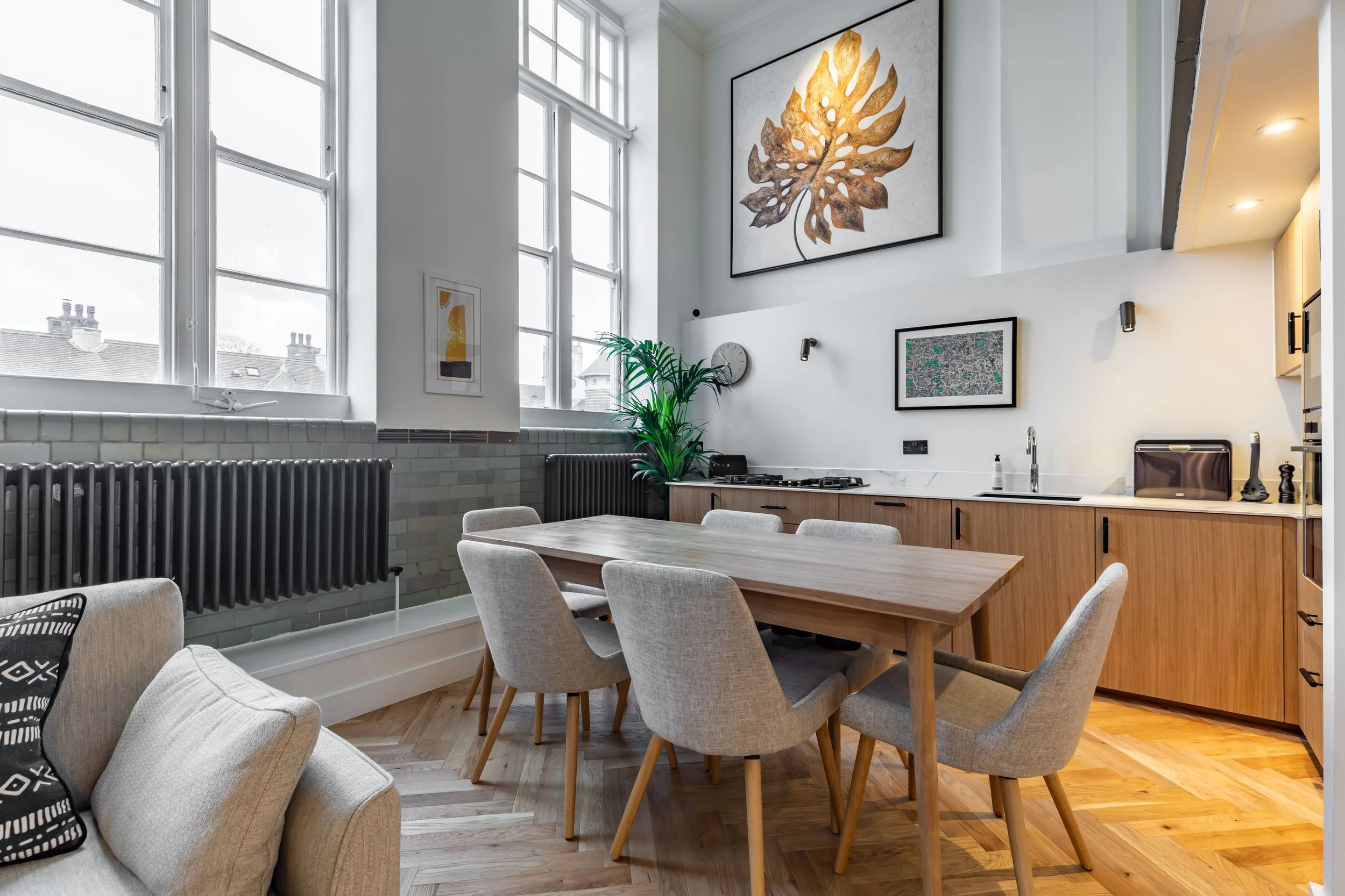 Dining area in a converted Grade II listed school duplex London — oak dining table and chairs, full-height original sash windows, glazed sage dado tiles, original column radiators, kitchen beyond, golden leaf artwork above. Studio Akiva