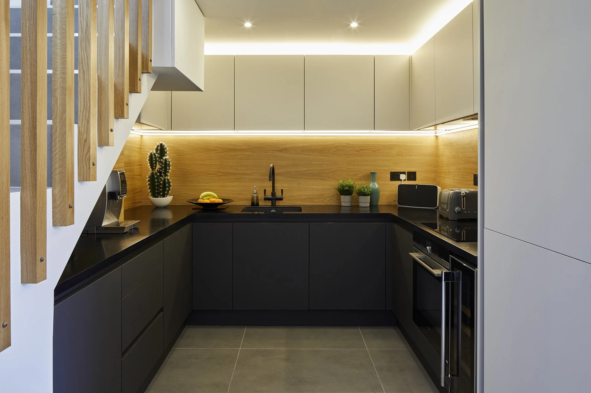 Industrial kitchen at night in a Shoreditch loft — LED strip lighting illuminates the oak splashback from beneath the pale upper cabinets, creating warm ambient light against the matte black kitchen. Studio Akiva, London