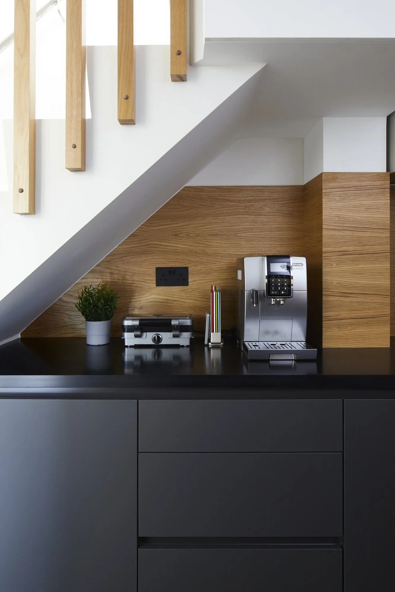 Under-stair kitchen counter zone in a Shoreditch loft — matte black handleless drawers, oak panel backdrop, black worktop and De'Longhi coffee machine. Clever use of dead space in an industrial kitchen. Studio Akiva, London