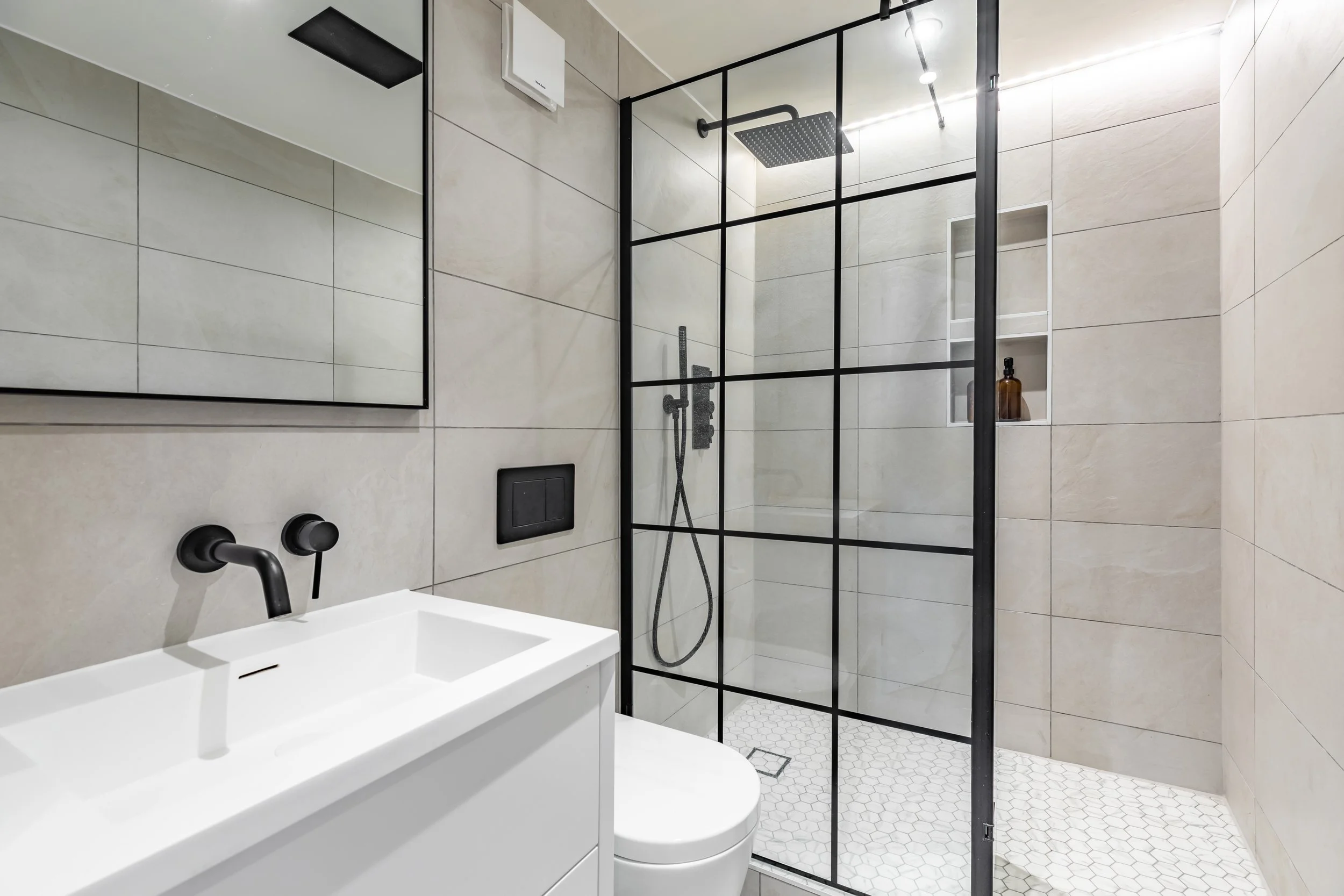 En-suite shower room in a school conversion duplex London — Crittall-style black-framed glass shower screen, matte black rain shower, pale stone tiles, hexagonal mosaic floor, wall niche, black-framed mirror cabinet. Studio Akiva