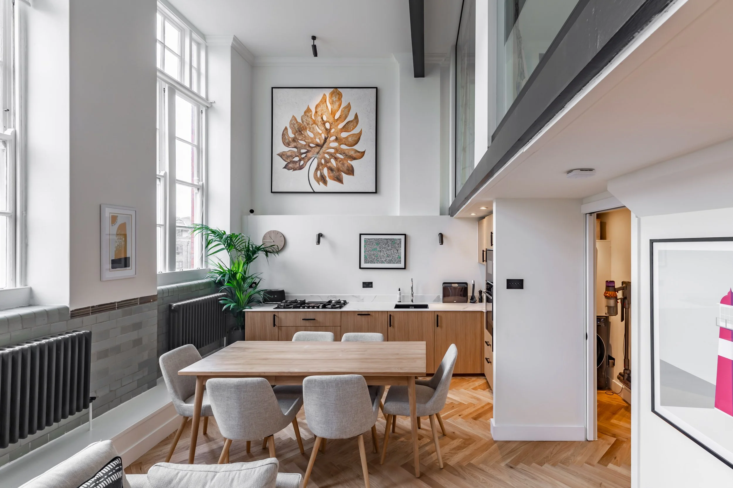 Kitchen and dining area in a converted boarding school duplex London — oak dining table, grey chairs, oak kitchen cabinets, white quartz worktop, double-height sash windows, original glazed dado tiles, mezzanine above. Interior design by Studio Akiva