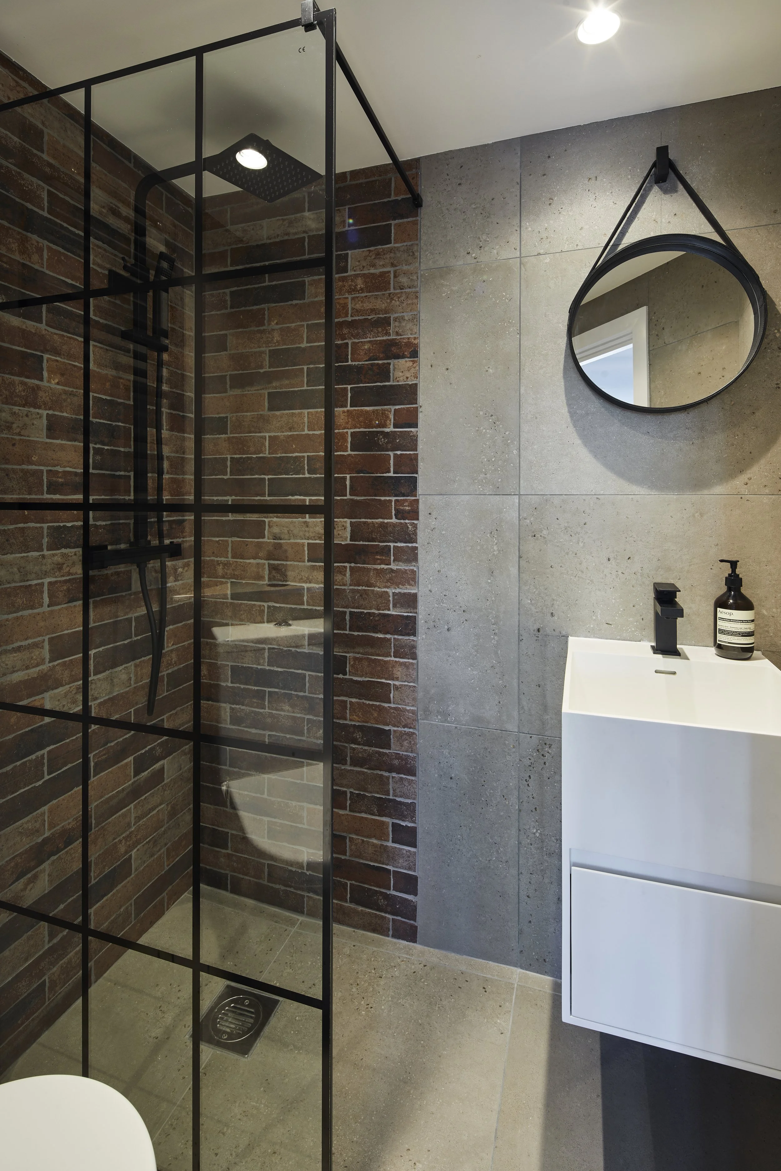 After photo — en-suite bathroom in a Shoreditch industrial loft with Crittall-style black-framed glass shower screen, exposed brick-effect tiles inside the shower, large-format concrete-effect grey wall tiles, round black-framed strap mirror, wall-hung vanity unit and matte black fittings. Interior design by Studio Akiva, London