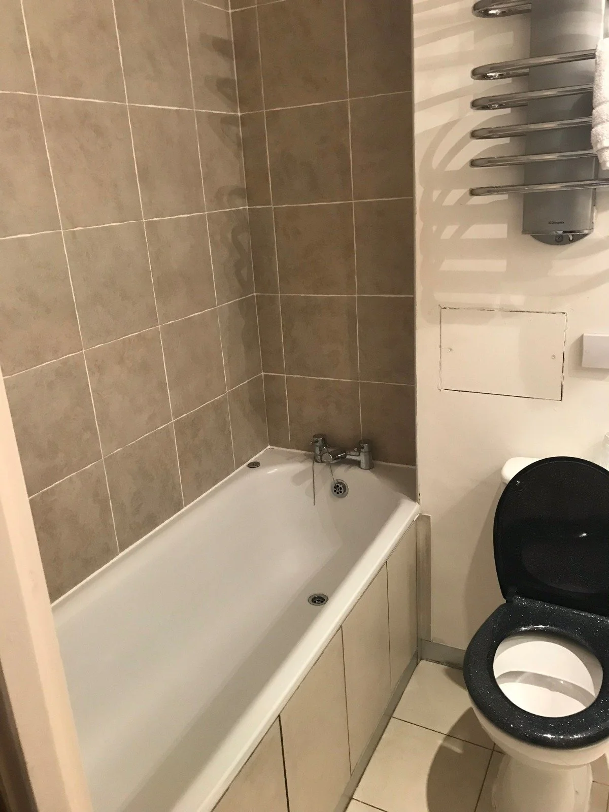 Before renovation photo of a main bathroom with beige tiles, white bathtub, chrome taps, a heated towel rail, and a mismatched black toilet seat