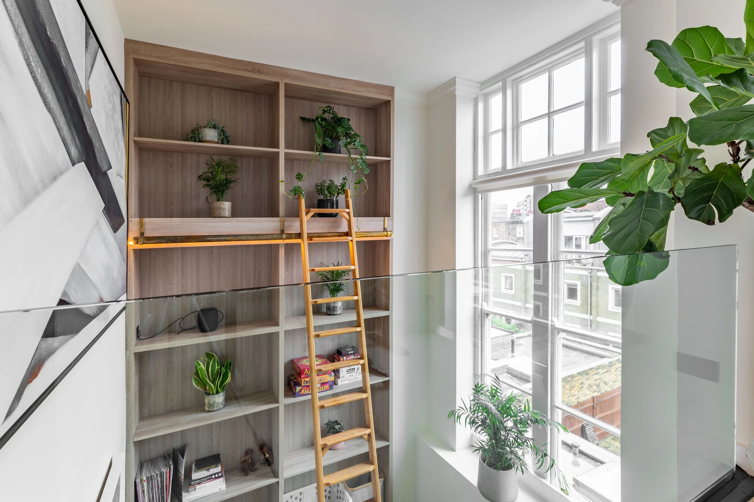 Mezzanine level of a converted boarding school duplex in London — bespoke full-height bookshelf with rolling library ladder, glass balustrade, fiddle leaf fig, original sash windows. Interior design by Studio Akiva
