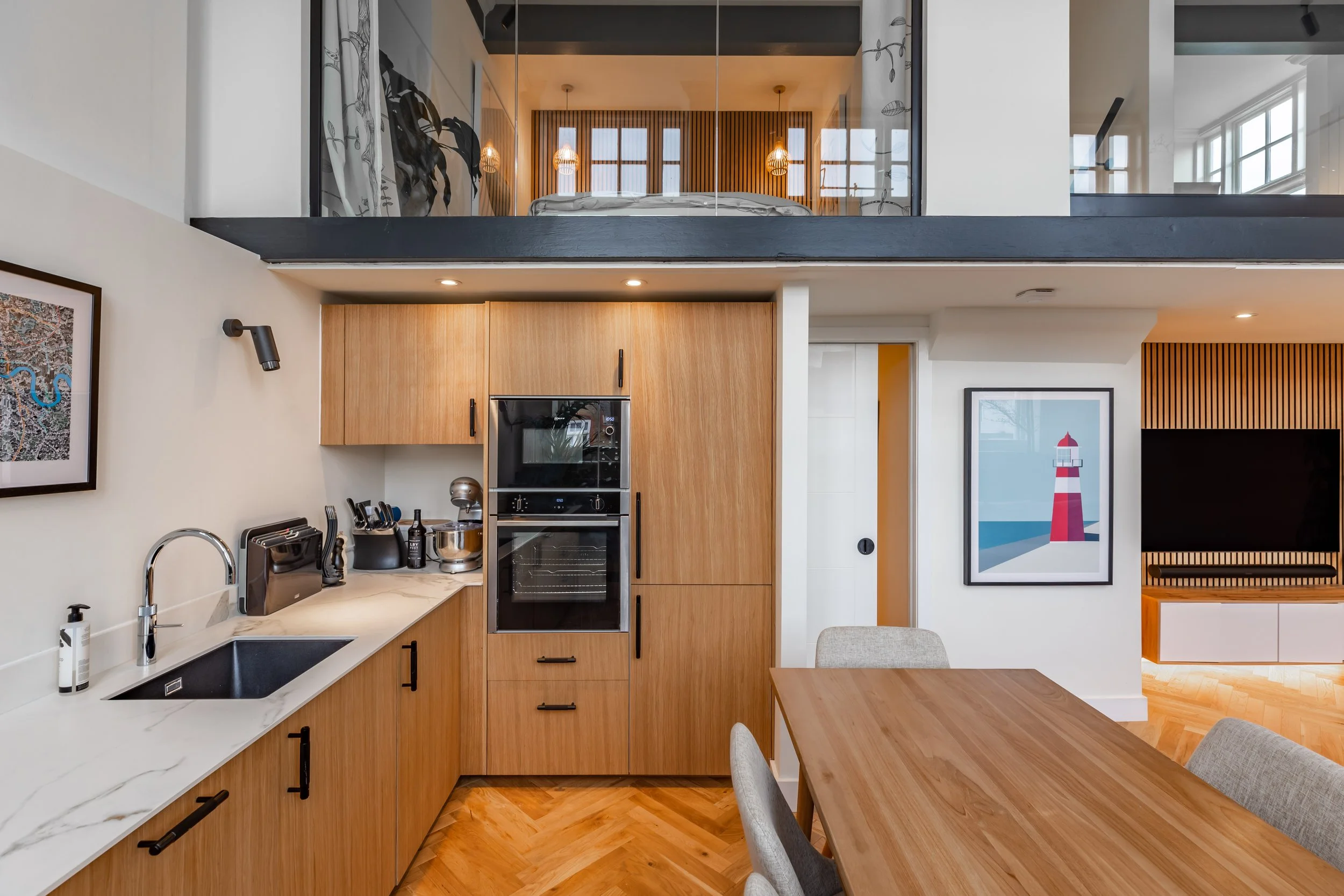 Kitchen detail in a Grade II listed school conversion duplex London — oak oven tower with Neff appliances, black undermount sink, Calacatta quartz worktop, black bar handles, herringbone floor, mezzanine bedroom warmly lit above through glass balustrade. Studio Akiva interior design