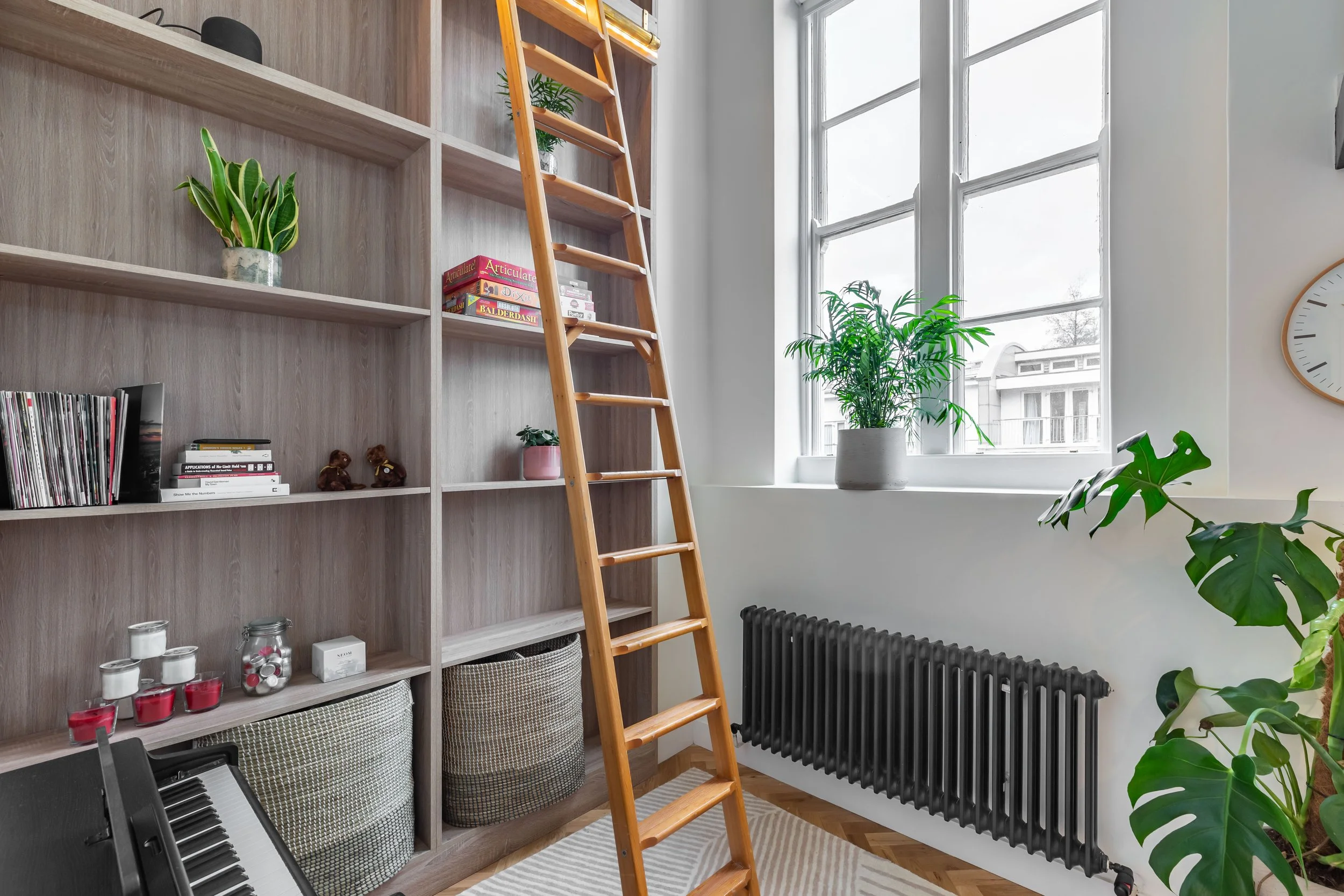 Bespoke full-height bookshelf with rolling library ladder in a Grade II listed London duplex — original tall sash windows, matte black column radiator, monstera plant, piano below. School conversion interior design by Studio Akiva