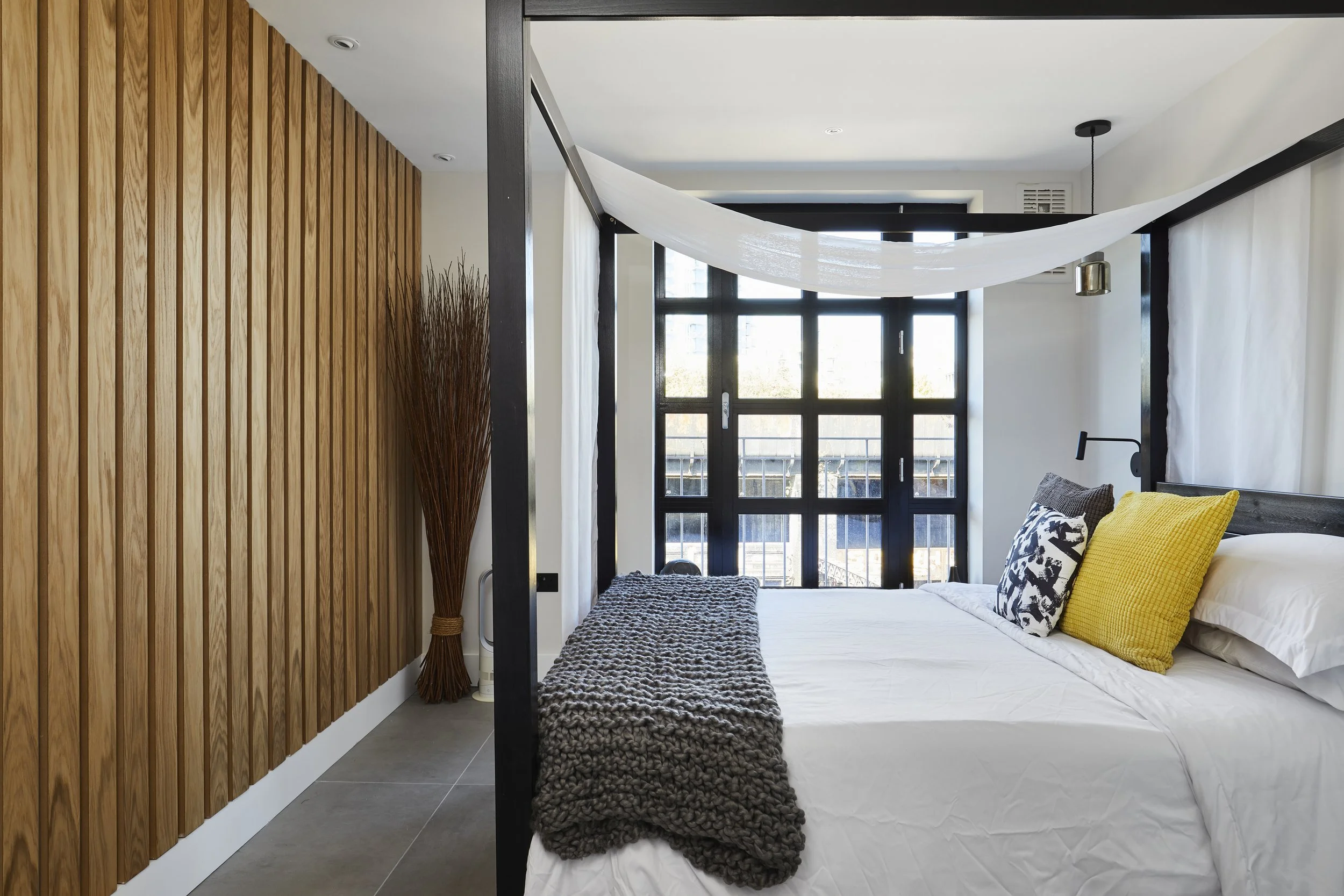 After photo — master bedroom in a Shoreditch industrial loft renovation with full-height oak slatted feature wall, matte black four-poster bed frame, black-framed grid balcony doors, large-format grey floor tiles, mustard and grey cushions, and a chunky knit throw. Interior design by Studio Akiva, London